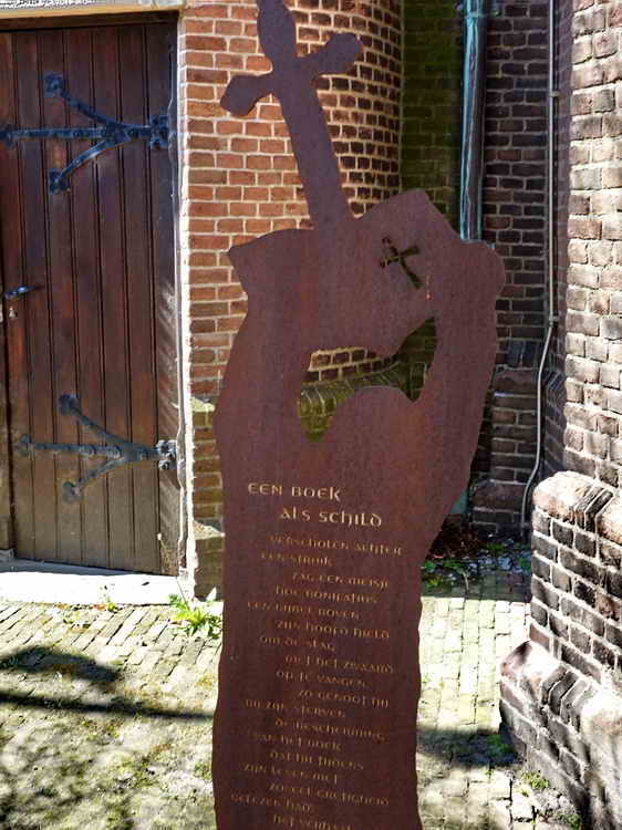 Dokkum, bij de Sint-Bonifatiuskerk, tekst: Een Boek als schild
Verscholen achter een struik zag een meisje hoe Bonifatius een Bijbel boven zijn hoofd hield om de slag met het zwaard op te vangen. Zo genoot hij bij zijn sterven de bescherming van het boek dat hij tijdens zijn leven met zoveel gretigheid gelezen had. Het verhaal werd doorverteld om duidelijk te maken dat Bonifatius over geen andere wapens wilde beschikken. Zijn enige wapen: het woord van God. Priesters mochten geen bloed vergieten. (22-4-2022)